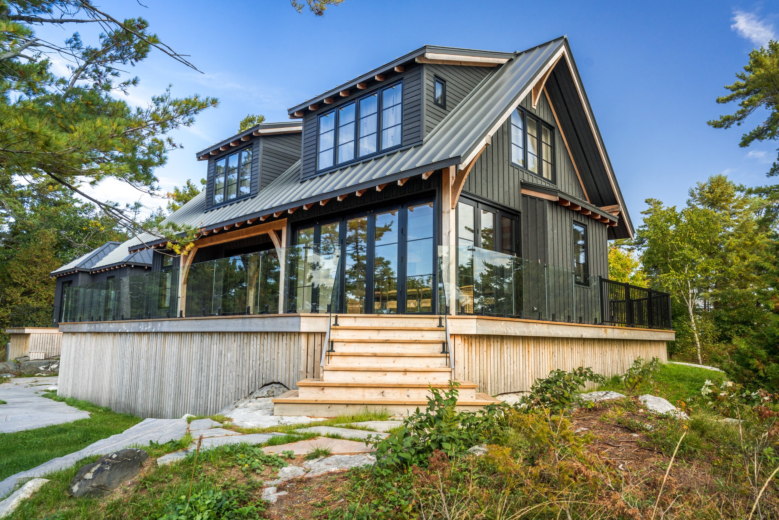 Modern two-story house with black wooden siding, large glass windows, and metal roofing, surrounded by trees and greenery.